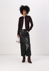 Svart cardigan over hvit topp, kombinert med et langt svart skinnskjørt med en frontspalte. Brune semskede støvler med hæl og en liten rosa veske.