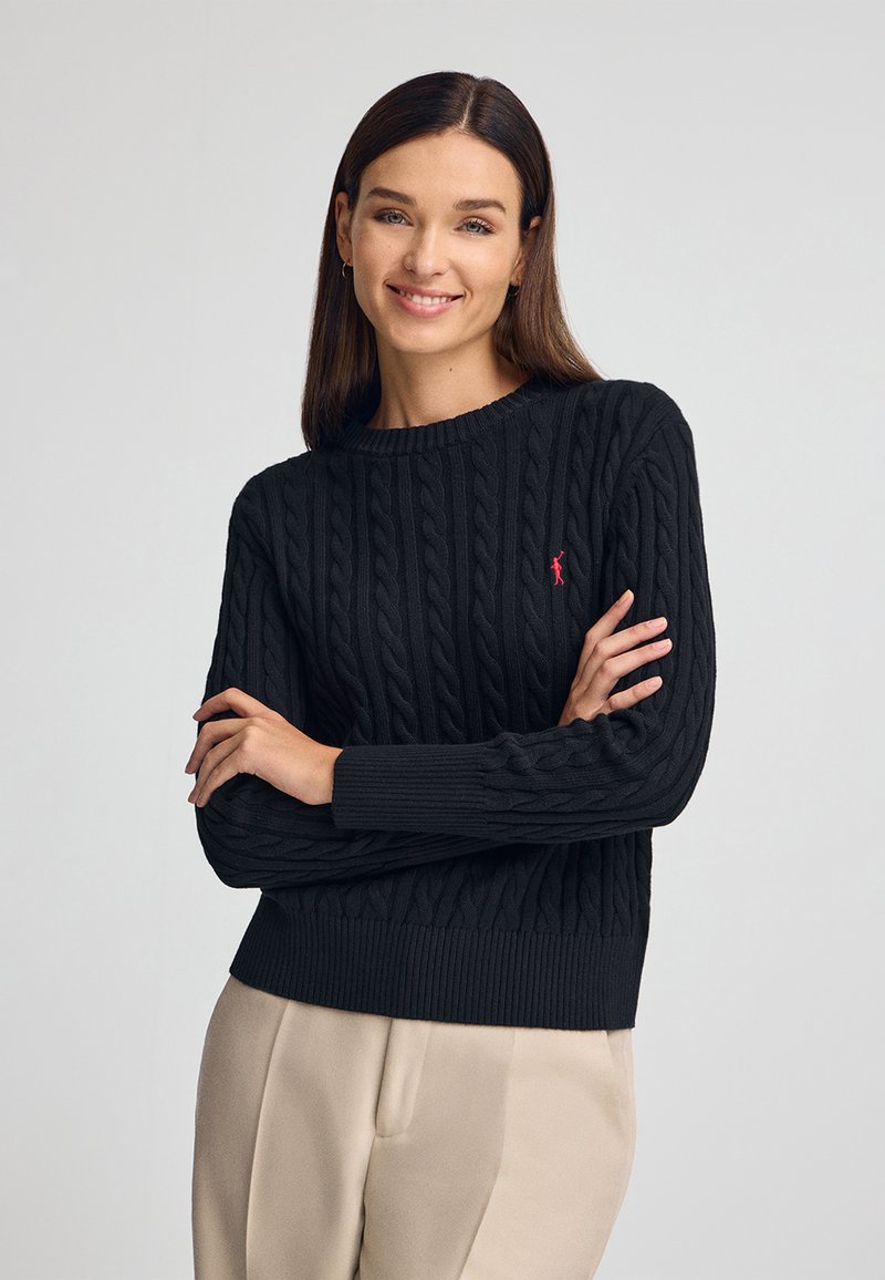 Pull en tricot noir à mailles avec un col rond et des poignets côtelés, arborant un petit logo rouge sur la poitrine gauche. Assorti à un pantalon beige.