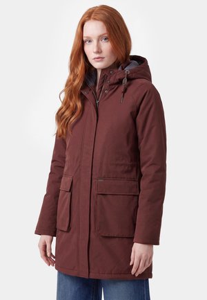 Giovane donna con lunghi capelli rossi che indossa un cappotto invernale marrone con cappuccio e grandi tasche frontali e jeans blu su sfondo semplice.