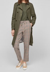 Trench coat verde oliva, top a coste verde scuro, pantaloni a quadri nei toni del rosa, beige e nero, abbinati a sneakers beige con lacci.