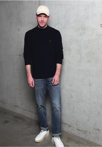 Un pull marin, un jean gris clair et des baskets blanches avec des accents verts. Porté avec une casquette beige clair, sur un fond en béton gris.