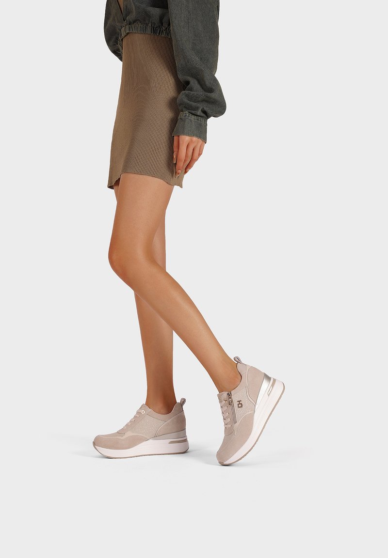 Scarpe da ginnastica color beige chiaro con tomaia in tessuto testurizzato, dettagli con lacci e cerniere, e suola bianca con un leggero tacco.