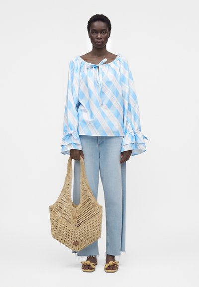 Femme portant un chemisier à carreaux bleu clair avec des manches évasées, un jean clair, des sandales jaunes et tenant un grand sac fourre-tout beige tricoté.