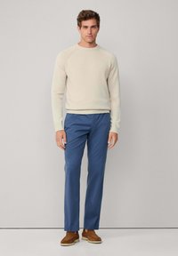 Suéter de punto beige claro con cuello redondo, combinado con pantalones azul ajustados. El modelo lleva puestas unas zapatos de ante marrón. Fondo liso.