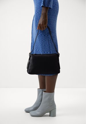 Borsa a spalla in pelle nera con una lunga tracolla, trama liscia e design minimalista, tenuta da una persona in un vestito blu a costine e stivali grigi.