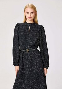 Robe noire à manches longues avec un décolleté en trou de serrure, motif à pois blancs, taille cintrée avec une ceinture noire et manches bouffantes.