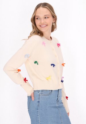 Béžový cardigan s knoflíkovým zapínáním, ozdobený barevnými látkovými mašlemi v různých odstínech, žebrovanými manžetami a zkráceným designem, v kombinaci s modrými džíny.
