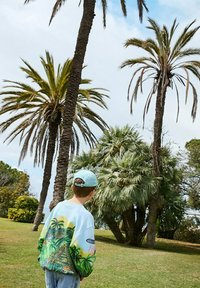 Felpa azzurra chiara con stampa tropicale vivace, caratterizzata da palme e una leopardo. Completa l'outfit un cappellino abbinato, jeans in denim e una vegetazione lussureggiante.