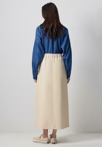 Denim long-sleeve shirt paired with a beige, knee-length skirt featuring an elastic waistband and a smooth texture, worn with beige sneakers.