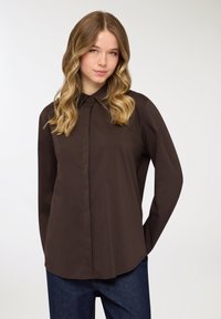 Camicia marrone a maniche lunghe con bottoni e colletto, caratterizzata da un orlo dritto e una texture liscia, abbinata a pantaloni di denim blu.