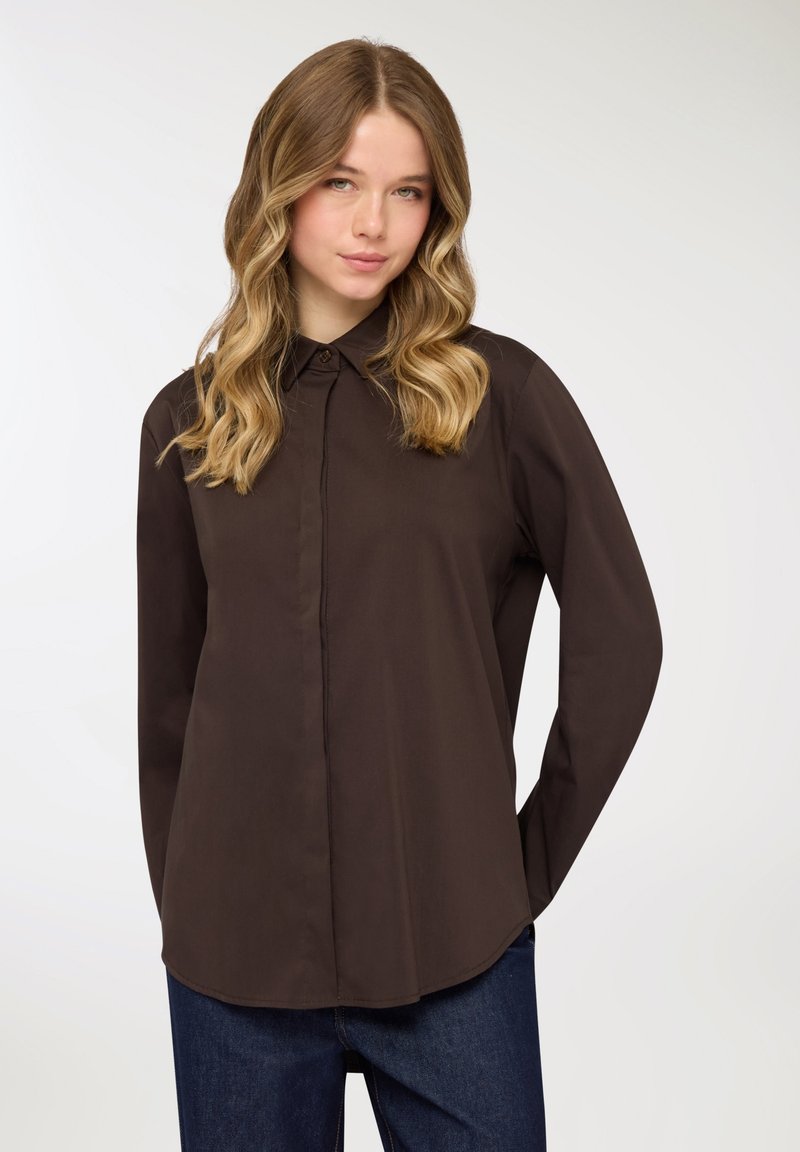 Camicia marrone a maniche lunghe con bottoni e colletto, caratterizzata da un orlo dritto e una texture liscia, abbinata a pantaloni di denim blu.