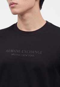 Νέος άνδρας φοράει μαύρο t-shirt Armani Exchange με ανάγλυφο κείμενο "Armani Exchange Milano / New York" στο στήθος.