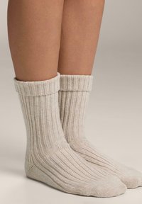 Nogi w kremowych, prążkowanych skarpetkach z podwiniętymi mankietami, stojące na neutralnej beżowej powierzchni.