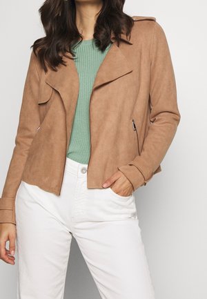 Femme portant une veste en daim beige par-dessus un top en maille verte et un pantalon blanc taille haute, avec une main dans la poche sur un fond uni.