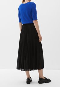 Person i en blue kortärmad topp, svart plisserad midi kjol och svarta loafers, står mot en vit bakgrund, vänd bort.