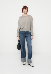 Lichtgrijze lange mouw top gecombineerd met hoog-was blauwe denim flare broeken met contrasterende boorden. Model houdt een zwarte tas vast.