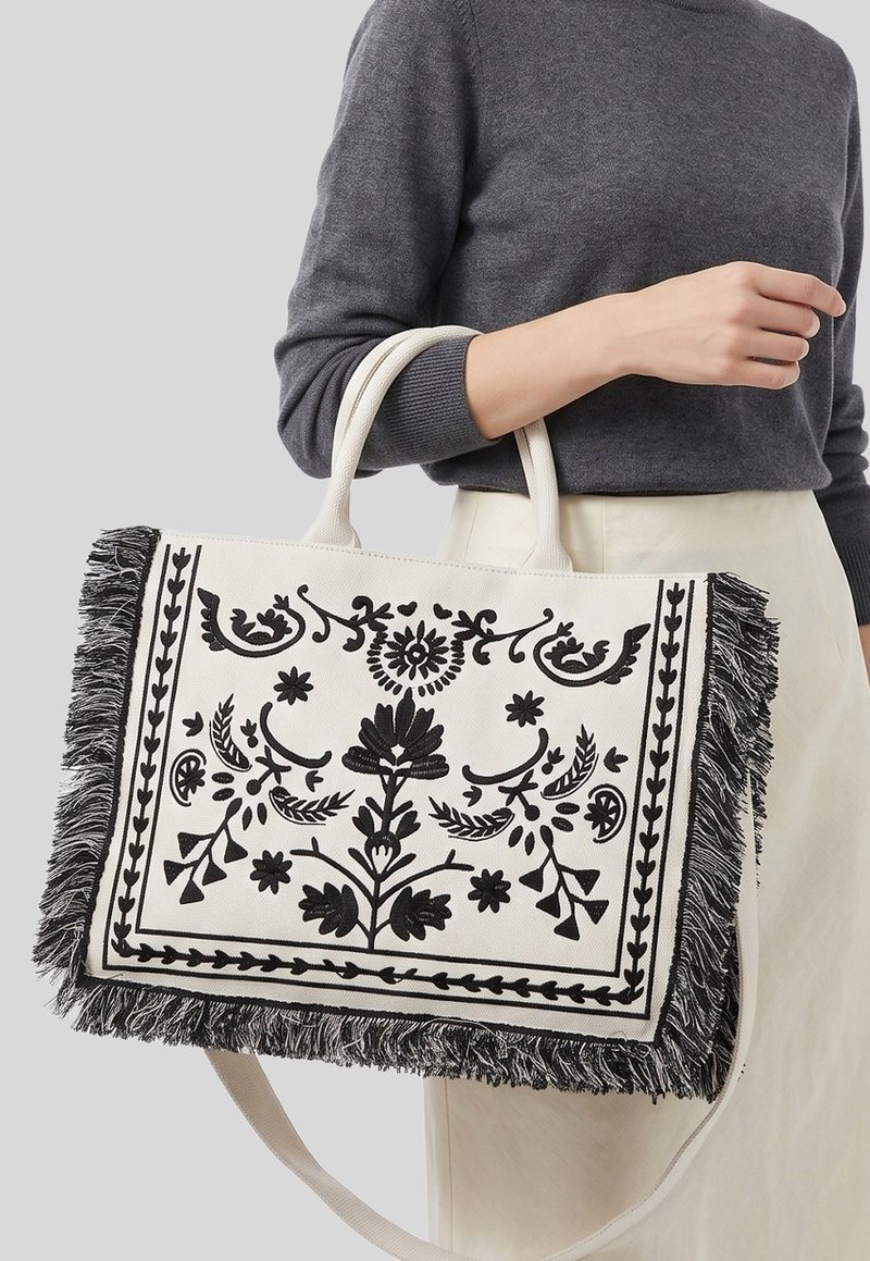 Femme en pull gris et jupe blanche tenant un grand sac cabas crème avec broderie florale noire et bords à franges.