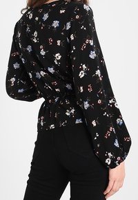 Blusa nera floreale con lunghe maniche a sbuffo, con vita cinchata. Il tessuto presenta piccoli fiori blu e bianchi su uno sfondo scuro.