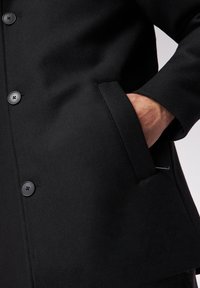 Zwarte wollen jas met een gladde textuur, knoopsluiting, zijzakken en een licht getailleerde pasvorm. Opvallende details zijn de zwarte knopen.