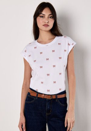 Le mannequin porte une chemise blanche avec de petits motifs de nœuds roses, associée à un jeans foncé et une ceinture marron, se tenant devant un fond uni.