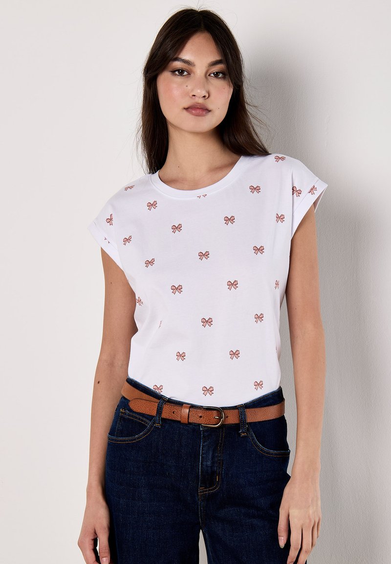 El modelo lleva una camisa blanca con pequeños patrones de lazo rosa, combinada con unos jeans oscuros y un cinturón marrón, de pie contra un fondo liso.