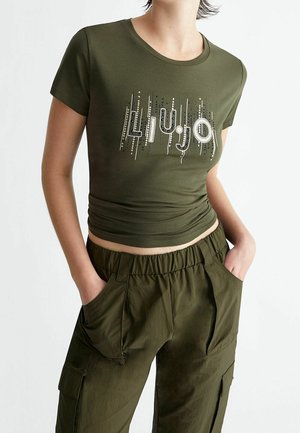 Osoba ubrana w dopasowany oliwkowozielony T-shirt z logo "LIU·JO" ozdobionym koralikami, w parze z luźnymi oliwkowozielonymi spodniami typu cargo, ręce w kieszeniach.