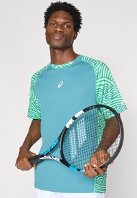 Τιρκουάζ κοντομάνικο μπλουζάκι τένις με πράσινα μανίκια με σχέδιο, διαθέτει λαιμόκοψη και λογότυπο στο στήθος. Το άτομο κρατά μια ρακέτα τένις.