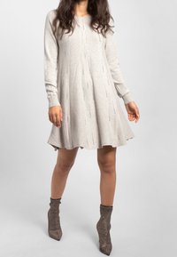 Femme portant une robe en tricot à manches longues beige clair avec des motifs verticaux subtils et des bottines à talon pointu étincelantes sur un fond uni.
