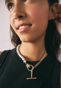 Zlatý náhrdelník s perlami, obsahujúci okrúhly prívesok so hviezdou, vrstvené reťaze a zapínanie na toggle, nosený cez čierny top.