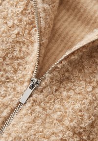 Gros plan d'une veste en laine beige à texture bouclée partiellement dézippée avec une fermeture éclair métallique argentée brillante.
