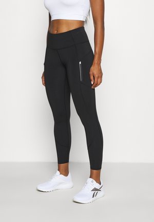 Athletisk person iført sorte højtaljede tights med sidelommer med lynlås og hvide løbesko, stående foran en hvid baggrund.