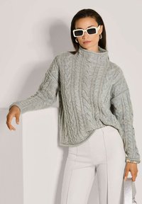 Frau trägt einen hellgrauen, grob gestrickten Rollkragenpullover, weiße Hosen, weiße rechteckige Sonnenbrille und hält eine silberne Handtasche.