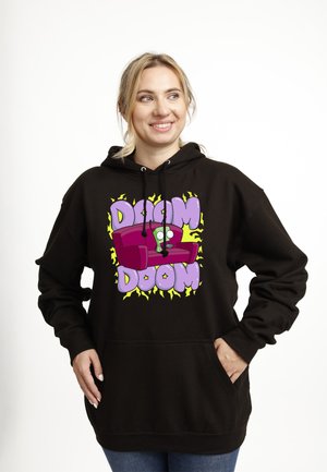 Femme souriante portant un sweat à capuche noir avec un personnage vert de dessin animé, assise sur un canapé rouge avec les mots « DOOM DOOM » en lettres violettes.