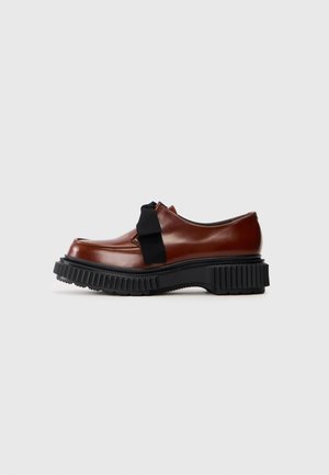 Mocassin en cuir marron avec semelle épaisse noire striée et un large nœud en tissu noir sur fond blanc.