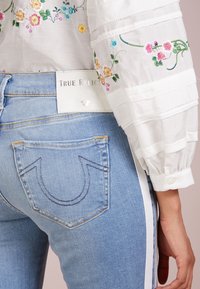 Jeans de denim azul claro con bolsillos traseros cosidos y franjas laterales blancas. Combinados con una blusa blanca bordada con patrones florales.