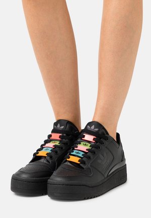 Black Adidas sneakers with thick soles, worn on bare legs, featuring colorful plastic lace clips in pink, green, blue, and orange on black laces.