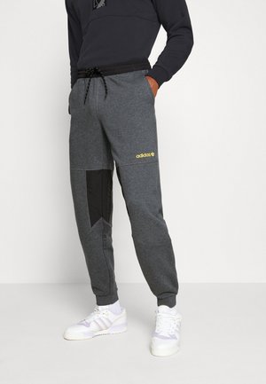 Persoon die donkere grijze Adidas joggers met zwarte accenten, een zwarte trui en witte sneakers draagt, staand tegen een effen witte achtergrond.