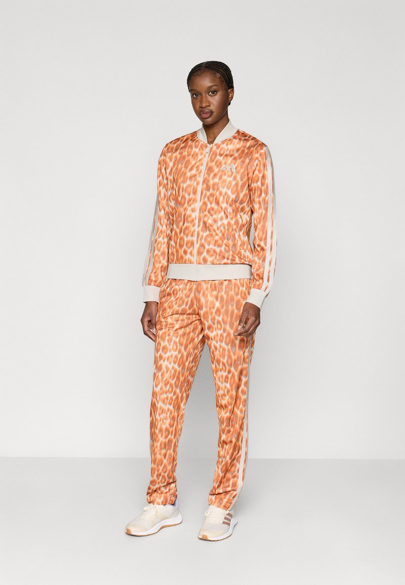 Oranžs sporta kostīms ar leoparda rakstu, ieskaitot rāvējslēdzēja jaku un atbilstošas bikses, ar baltām svītrām gar piedurknēm un kājām.