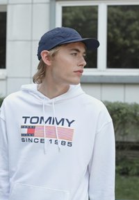 Fehér kapucnis pulcsi "TOMMY" logóval és "SINCE 1985" felirattal tengerészkék és piros színben. Tengerész sapka és világos háttér z�öld növényzettel és ablakokkal.