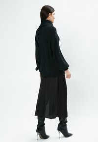 Schwarzer Strickpullover mit hohem Kragen, lockerer Passform und herunterhängenden Schultern, kombiniert mit einem langen schwarzen Rock und knielangen schwarzen Stiefeln mit Absatz.
