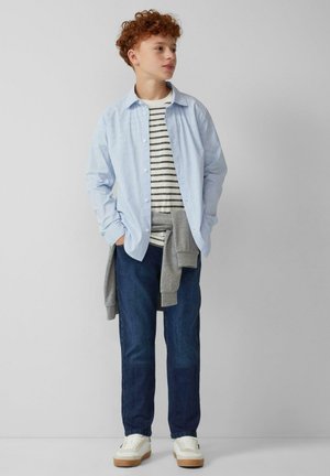 Lichtblauw gestreept overhemd over een grijs gestreept T-shirt, gecombineerd met donkere jeans en witte sneakers. Informele outfit met gelaagde texturen.