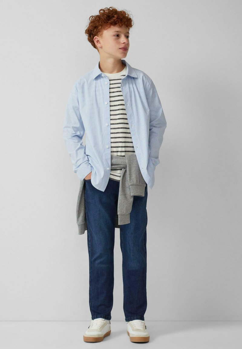 Lichtblauw gestreept overhemd over een grijs gestreept T-shirt, gecombineerd met donkere jeans en witte sneakers. Informele outfit met gelaagde texturen.
