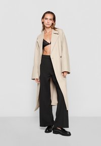 Beige long coat over a black lace bralette, paired with high-waisted black wide-leg pants and black lace-up shoes. Smooth texture, tailored design.