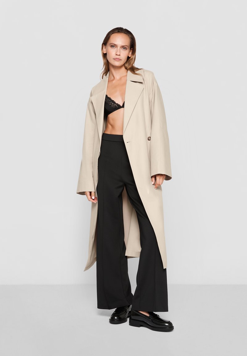 Beige long coat over a black lace bralette, paired with high-waisted black wide-leg pants and black lace-up shoes. Smooth texture, tailored design.