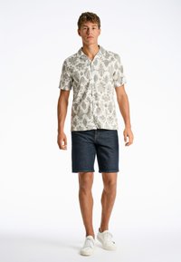 Camisa de manga corta con un fondo beige claro y un patrón floral verde oscuro, combinada con pantalones cortos de mezclilla oscura y zapatillas deportivas blancas.