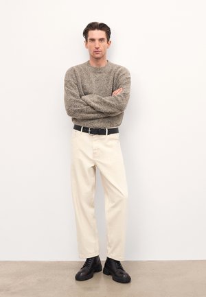 Pantalones de algodón beige claro combinados con un suéter marrón texturizado y botas negras con cordones. El modelo está de pie con los brazos cruzados frente a un fondo liso.