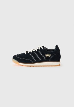 Sort Adidas SL 72 sneaker med hvide snørebånd, tre hvide striber, gult Adidas-logo og lysebrun sål mod en ensfarvet baggrund.