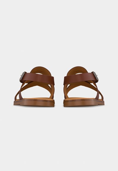 Ζευγάρι καφέ δερμάτινων σανδαλιών με λουράκια στον αστράγαλο και ασημένιες αγκράφες, εμφανίζονται από πίσω σε απλό ανοιχτό φόντο.