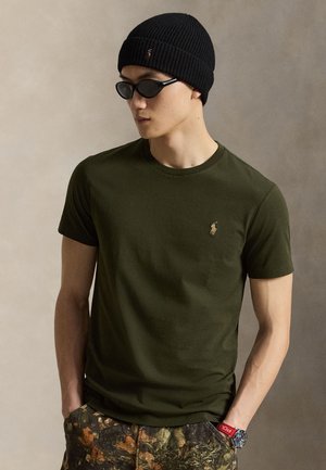 Mann trägt schwarze Beanie, schwarze Sonnenbrille, olivgrünes T-Shirt mit kleinem Logo, Tarnhose und eine rote Armbanduhr, steht mit Händen in den Taschen.
