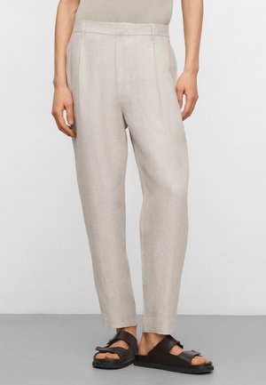 Pantaloni beige chiaro su misura in lino indossati con sandali a punta aperta marrone scuro, mostrati dalla vita ai piedi su uno sfondo neutro.